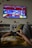 A person is lounging on a couch, holding a Sony game controller, playing a video game displayed on a wall-mounted TV screen. The game has bright and colorful graphics with red and blue tones. The room includes decorative elements like shelves, small potted plants, and wall-mounted ornaments.