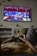 A person is lounging on a couch, holding a Sony game controller, playing a video game displayed on a wall-mounted TV screen. The game has bright and colorful graphics with red and blue tones. The room includes decorative elements like shelves, small potted plants, and wall-mounted ornaments.