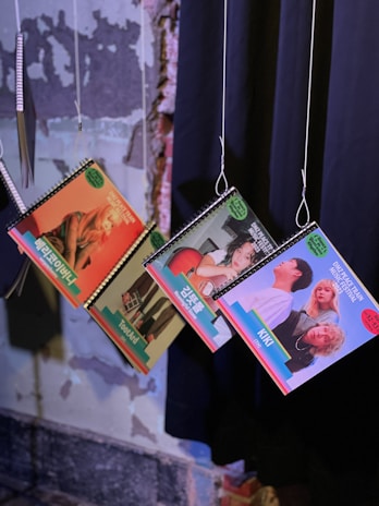 Three spiral-bound booklets with colorful covers are hanging from strings against a textured wall. Each cover features different people and text in a language that appears to be Korean.