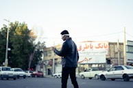 A person wearing headphones is standing at an intersection, looking down at a mobile device. Several cars are on the road, and there are buildings in the background with signs in Persian script. The scene is set during the day with a clear sky.