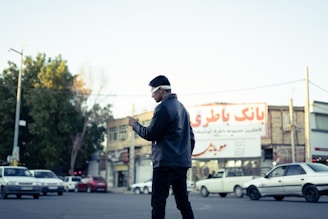 A person wearing headphones is standing at an intersection, looking down at a mobile device. Several cars are on the road, and there are buildings in the background with signs in Persian script. The scene is set during the day with a clear sky.