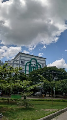 A multi-story building with a distinctive green design on the facade is surrounded by lush trees and a well-maintained grassy area. The sky is partly cloudy, with a mix of large dark clouds and blue sky showing through.