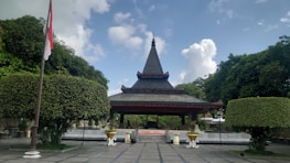 A traditional pavilion with a pointed roof stands prominently in a landscaped area, surrounded by lush green trees. The structure is elevated on a set of steps and is flanked by neatly trimmed hedges shaped into spherical forms, with an Indonesian flag visible on the left side. The sky is bright with scattered clouds, creating a serene outdoor atmosphere.