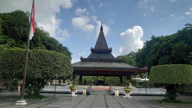 A traditional pavilion with a pointed roof stands prominently in a landscaped area, surrounded by lush green trees. The structure is elevated on a set of steps and is flanked by neatly trimmed hedges shaped into spherical forms, with an Indonesian flag visible on the left side. The sky is bright with scattered clouds, creating a serene outdoor atmosphere.
