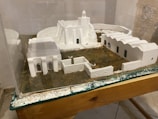 A miniature model of a building complex featuring white structures resembling traditional architectural styles. The model includes several interconnected buildings with arched doorways and windows, all set on a textured grassy base. Protective glass encases the display, which rests on a wooden surface.