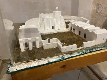 A miniature model of a building complex featuring white structures resembling traditional architectural styles. The model includes several interconnected buildings with arched doorways and windows, all set on a textured grassy base. Protective glass encases the display, which rests on a wooden surface.