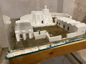 A miniature model of a building complex featuring white structures resembling traditional architectural styles. The model includes several interconnected buildings with arched doorways and windows, all set on a textured grassy base. Protective glass encases the display, which rests on a wooden surface.