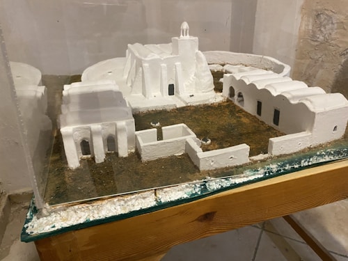 A miniature model of a building complex featuring white structures resembling traditional architectural styles. The model includes several interconnected buildings with arched doorways and windows, all set on a textured grassy base. Protective glass encases the display, which rests on a wooden surface.