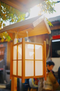 A traditional wooden lantern with a rectangular shape and white panels is hanging under a roof-like structure. The lantern is illuminated by sunlight and surrounded by green foliage. In the background, there is a blurred scene with people and a building, adding to a cozy and rustic atmosphere.