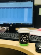 A desk featuring a computer monitor with a blurred spreadsheet visible, a keyboard, and a cup of coffee with a black and green sleeve and lid, sitting on a wooden desk. A black computer mouse is also present beside the coffee cup.