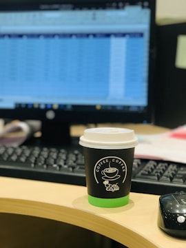 A desk featuring a computer monitor with a blurred spreadsheet visible, a keyboard, and a cup of coffee with a black and green sleeve and lid, sitting on a wooden desk. A black computer mouse is also present beside the coffee cup.