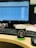 A desk featuring a computer monitor with a blurred spreadsheet visible, a keyboard, and a cup of coffee with a black and green sleeve and lid, sitting on a wooden desk. A black computer mouse is also present beside the coffee cup.