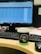 A desk featuring a computer monitor with a blurred spreadsheet visible, a keyboard, and a cup of coffee with a black and green sleeve and lid, sitting on a wooden desk. A black computer mouse is also present beside the coffee cup.