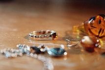 A collection of jewelry items is arranged on a wooden surface. The centerpiece is a gold ring with a series of links and a central amber stone. Nearby, a delicate chain and earrings can be seen, along with a glass figurine, possibly with an etched design, in a warm, amber tone.