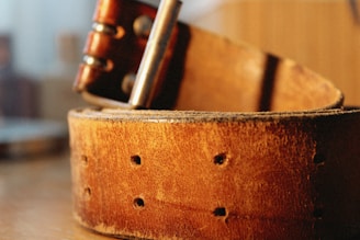 A close-up view of a well-worn brown leather belt with visible holes and a metal buckle. The texture of the leather is rough and aged, with subtle variations in color and scuffs visible. The metal elements are slightly tarnished, enhancing the vintage appearance.
