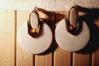 A pair of round, white earrings with a gold accent on top is placed on a wooden surface. The earrings have an elegant design with a shiny texture.
