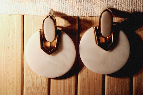 A pair of round, white earrings with a gold accent on top is placed on a wooden surface. The earrings have an elegant design with a shiny texture.