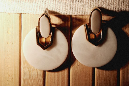 A pair of round, white earrings with a gold accent on top is placed on a wooden surface. The earrings have an elegant design with a shiny texture.