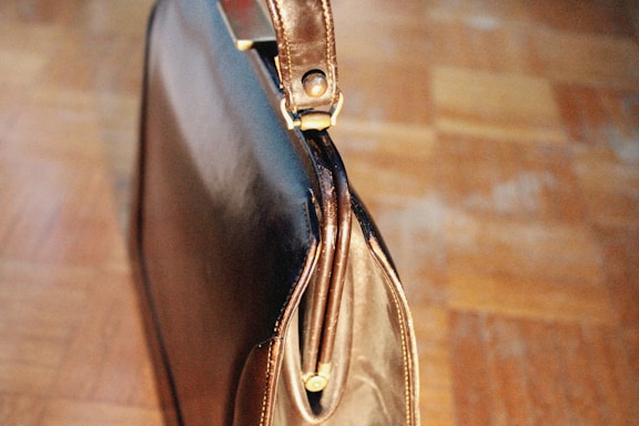 A leather briefcase with a shiny surface and a brass clasp is placed on a wooden floor. The briefcase is slightly open, revealing a zipper and intricate stitching details.