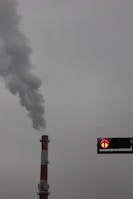 A tall industrial chimney emits a thick plume of smoke into the overcast sky. To the right, a traffic signal displays a red arrow pointing upwards.