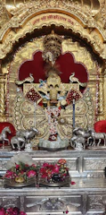 An ornate shrine with a richly decorated idol adorned with detailed golden patterns and intricate jewelry. The idol is set against a backdrop of gold and red elements. There are statues of horses on both sides, and offerings such as flowers and monetary notes are placed in front.