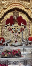 An ornate shrine with a richly decorated idol adorned with detailed golden patterns and intricate jewelry. The idol is set against a backdrop of gold and red elements. There are statues of horses on both sides, and offerings such as flowers and monetary notes are placed in front.