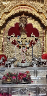 An ornate shrine with a richly decorated idol adorned with detailed golden patterns and intricate jewelry. The idol is set against a backdrop of gold and red elements. There are statues of horses on both sides, and offerings such as flowers and monetary notes are placed in front.