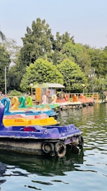 A row of colorful paddle boats shaped like swans is lined up on a serene body of water. The boats are in various colors, including purple, yellow, and orange, and are numbered. In the background, a lush green park with dense trees can be seen. There is a small dock with a sign indicating paddle boating, and a person is visible near the dock.