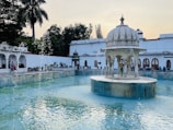 An ornate white pavilion with arches sits at the center of a tranquil, light blue pool surrounded by greenery and historical architecture. The water gently flows from the decorative elements of the pavilion. People are casually seated around the perimeter of the pool, enjoying the serene environment under a clear sky, with palm trees on one side.