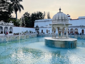 An ornate white pavilion with arches sits at the center of a tranquil, light blue pool surrounded by greenery and historical architecture. The water gently flows from the decorative elements of the pavilion. People are casually seated around the perimeter of the pool, enjoying the serene environment under a clear sky, with palm trees on one side.