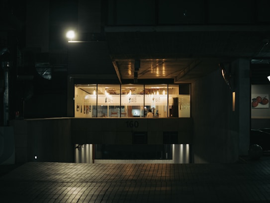 A dimly lit office building exterior at night, with a warm glow coming from inside the glass-paneled windows. A person is visible seated at a desk, suggesting a late night work environment. The entrance and surroundings are dark, illuminated by a solitary street lamp above.