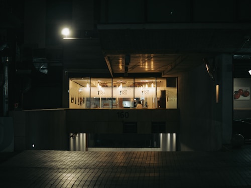 A dimly lit office building exterior at night, with a warm glow coming from inside the glass-paneled windows. A person is visible seated at a desk, suggesting a late night work environment. The entrance and surroundings are dark, illuminated by a solitary street lamp above.