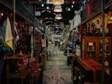 A bustling indoor market features an array of colorful textiles, clothing, and various antiques. The aisles are narrow with items displayed on both sides, and the ceiling is adorned with hanging lights. The market has a rustic and eclectic ambiance, with intricate wooden furniture and vibrant fabric patterns adding to the visual richness.