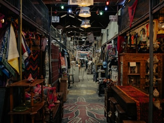A bustling indoor market features an array of colorful textiles, clothing, and various antiques. The aisles are narrow with items displayed on both sides, and the ceiling is adorned with hanging lights. The market has a rustic and eclectic ambiance, with intricate wooden furniture and vibrant fabric patterns adding to the visual richness.