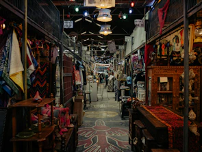A bustling indoor market features an array of colorful textiles, clothing, and various antiques. The aisles are narrow with items displayed on both sides, and the ceiling is adorned with hanging lights. The market has a rustic and eclectic ambiance, with intricate wooden furniture and vibrant fabric patterns adding to the visual richness.