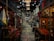 A bustling indoor market features an array of colorful textiles, clothing, and various antiques. The aisles are narrow with items displayed on both sides, and the ceiling is adorned with hanging lights. The market has a rustic and eclectic ambiance, with intricate wooden furniture and vibrant fabric patterns adding to the visual richness.