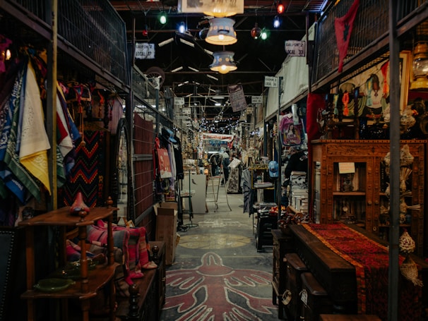 A bustling indoor market features an array of colorful textiles, clothing, and various antiques. The aisles are narrow with items displayed on both sides, and the ceiling is adorned with hanging lights. The market has a rustic and eclectic ambiance, with intricate wooden furniture and vibrant fabric patterns adding to the visual richness.