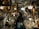 A person stands in a cluttered antique store filled with various vintage items. The shelves and walls are adorned with ornate picture frames, decorative plates, and glassware. Chandeliers and lamps hang from the ceiling, casting a warm glow over the scene.