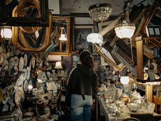 A person stands in a cluttered antique store filled with various vintage items. The shelves and walls are adorned with ornate picture frames, decorative plates, and glassware. Chandeliers and lamps hang from the ceiling, casting a warm glow over the scene.