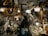 A person stands in a cluttered antique store filled with various vintage items. The shelves and walls are adorned with ornate picture frames, decorative plates, and glassware. Chandeliers and lamps hang from the ceiling, casting a warm glow over the scene.