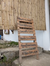 A wooden signboard is propped against a bamboo wall. The sign lists offerings such as vegetarian/vegan food, South Indian fare, speciality coffee, juices, smoothies, hemp experiments, and displays the hours of operation from 9 AM to 10 PM daily.