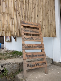 A wooden signboard is propped against a bamboo wall. The sign lists offerings such as vegetarian/vegan food, South Indian fare, speciality coffee, juices, smoothies, hemp experiments, and displays the hours of operation from 9 AM to 10 PM daily.