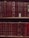 A bookshelf filled with rows of leather-bound books featuring intricate gold patterns and lettering on their spines. The books are mostly deep red in color, with a few exceptions of lighter red and brown shades. Some books have noticeable signs of wear, such as peeling labels or exposed sections where the leather has worn away, adding a vintage feel to the collection. A gold ribbon is visible sticking out of one of the books.