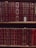 A bookshelf filled with rows of leather-bound books featuring intricate gold patterns and lettering on their spines. The books are mostly deep red in color, with a few exceptions of lighter red and brown shades. Some books have noticeable signs of wear, such as peeling labels or exposed sections where the leather has worn away, adding a vintage feel to the collection. A gold ribbon is visible sticking out of one of the books.