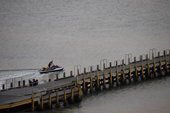 A person is riding a jet ski near a pier that extends into the water. The pier is long with numerous vertical supports and yellow safety railings. The water is calm, and there are a few people sitting on the edge of the pier.