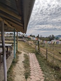 A patio area covers a pathway made of curved tiles leading to an open field. On the left, there are wooden tables and chairs under a roof with exposed beams. To the right, colorful prayer flags are strung along a wire fence, and the sky above is filled with fluffy clouds. The landscape includes patches of grass and distant hills.