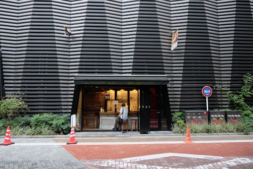 A small, modern kiosk with a stylish facade is set against a large building with a striking black and grey geometric pattern. The kiosk has an open counter with two stools and a person standing at the counter. Surrounding the kiosk are green plants, orange traffic cones, and a no parking sign. The street is paved with a mix of grey and red bricks.