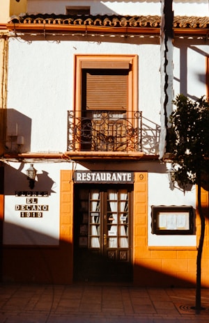 A small restaurant with a classic European facade featuring a sign reading 'Restaurante'. The building has white walls and orange trims, including a window with closed brown shutters and an iron balcony above the entry. The entrance door has glass panes with curtains behind them. A small black lamp is mounted on the wall beside an old plaque that reads 'Flores El Decano 1919'. Shadows cover part of the wall, and a tree partially obscures the view on the right.