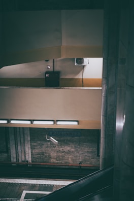 An indoor scene in a subway station with multiple levels of platforms. The walls are mostly beige with some yellow accents, and there are several light fixtures providing illumination. An air conditioning unit is attached to the wall. There is a sign indicating the station name, Piatra Unirii 2. An escalator is partially visible on the right side.