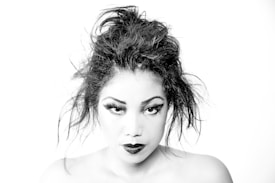 A black and white portrait of a person with an intense expression, featuring bold eyeliner and dark lipstick. The hair is styled in a messy, voluminous updo which adds to the dramatic look. The stark contrast enhances facial features and emphasizes the mood.